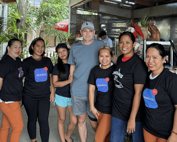 Group of people, including a man in a gray shirt, standing outdoors with a building and greenery in the background.